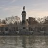 Equestrian statue on a tall pedestal inside a semicircular colonnade along a lake at sunset.