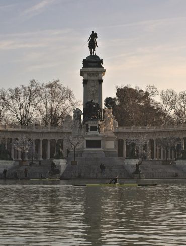 Equestrian statue on a tall pedestal inside a semicircular colonnade along a lake at sunset.