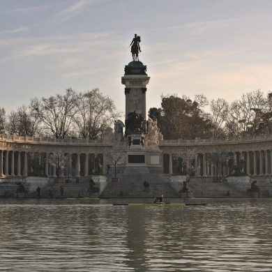 Equestrian statue on a tall pedestal inside a semicircular colonnade along a lake at sunset.