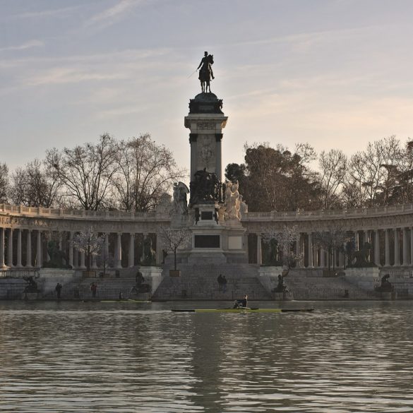 Equestrian statue on a tall pedestal inside a semicircular colonnade along a lake at sunset.