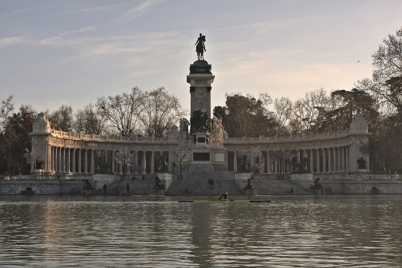 Equestrian statue on a tall pedestal inside a semicircular colonnade along a lake at sunset.