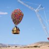 A colorful house lifted into the air by hundreds of balloons attached to a tall crane in a desert landscape