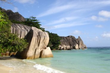 Tropical beach with large smooth granite boulders along the shoreline, turquoise sea and blue sky with wispy clouds.