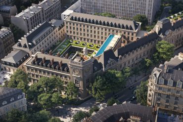 Aerial view of a historic city block with a central courtyard, blue rooftop pool, and manicured gardens.