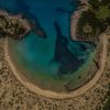 Aerial view of a crescent beach rimmed by rocky cliffs and turquoise water.