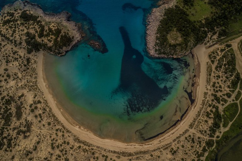 Aerial view of a crescent beach rimmed by rocky cliffs and turquoise water.