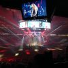 Crowd in a dark arena watching a live concert; a circular stage with musicians and large video screens overhead.
