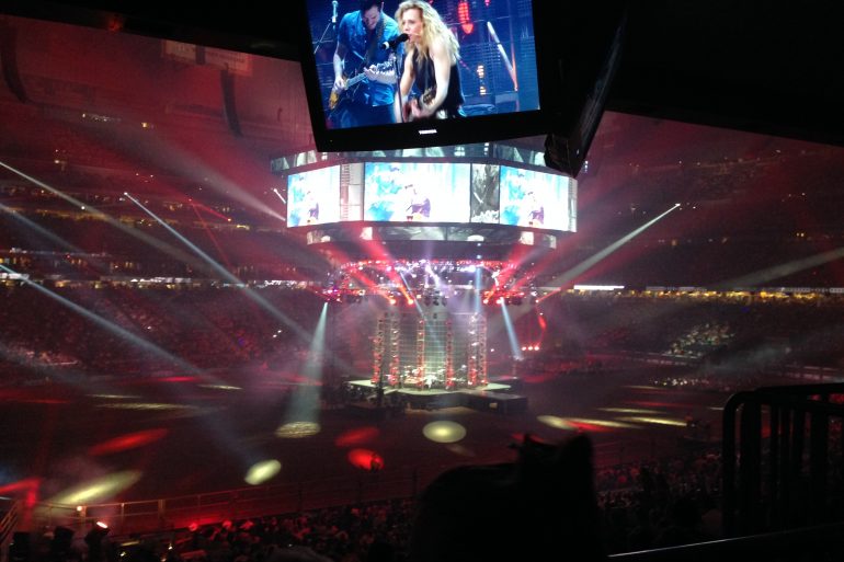 Crowd in a dark arena watching a live concert; a circular stage with musicians and large video screens overhead.