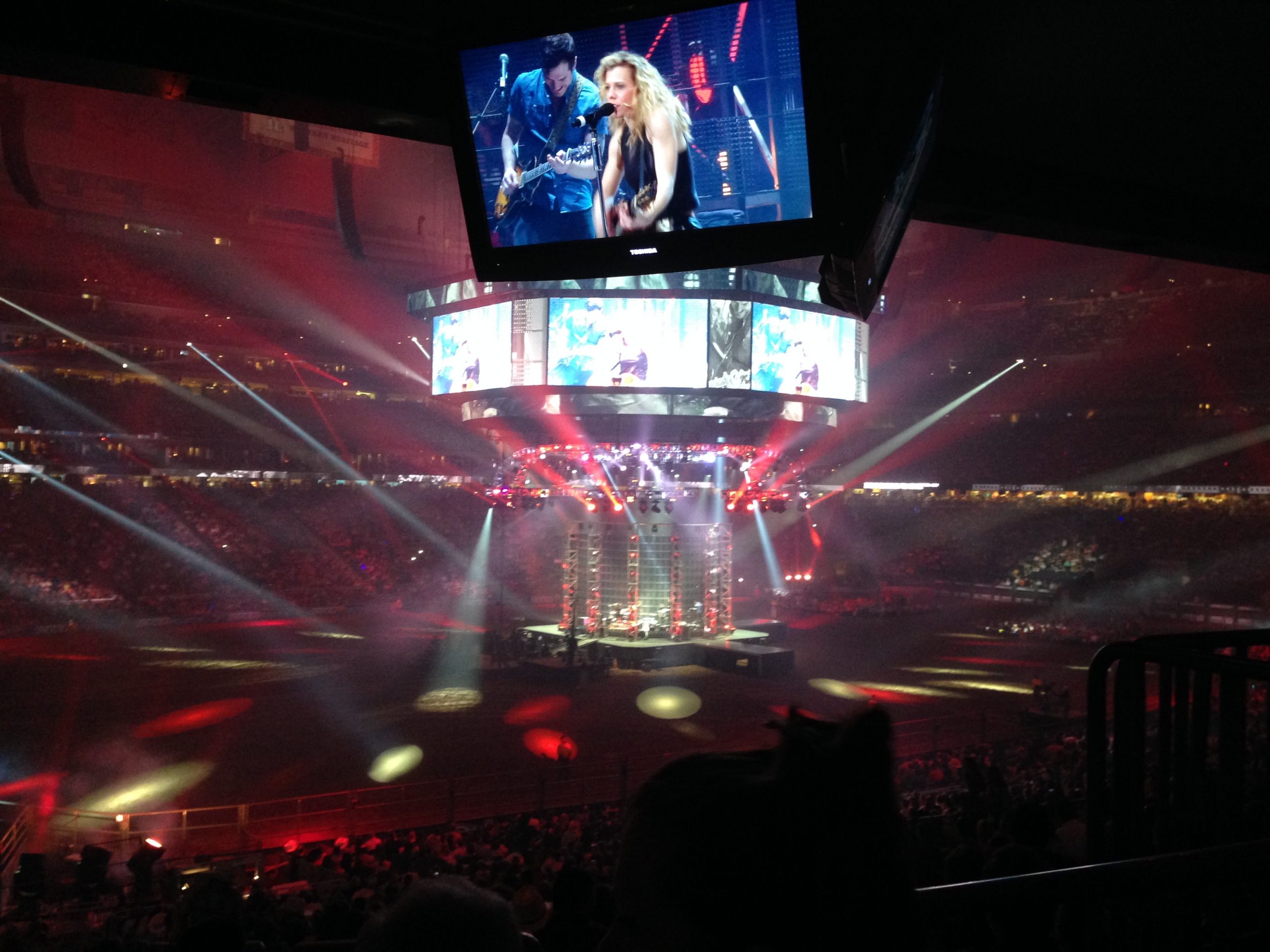 Crowd in a dark arena watching a live concert; a circular stage with musicians and large video screens overhead.