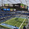 Circular video board hovering over the football field in SoFi Stadium, displaying team logos and game footage with empty blue seats around.