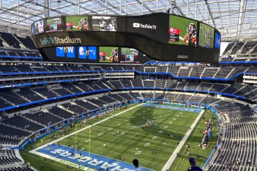 Circular video board hovering over the football field in SoFi Stadium, displaying team logos and game footage with empty blue seats around.