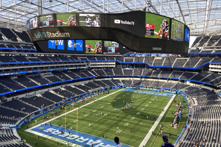 Circular video board hovering over the football field in SoFi Stadium, displaying team logos and game footage with empty blue seats around.