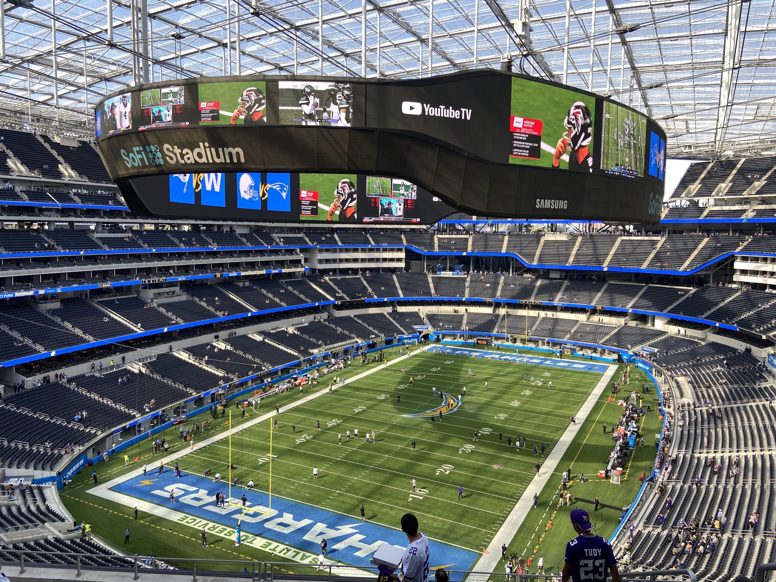 Circular video board hovering over the football field in SoFi Stadium, displaying team logos and game footage with empty blue seats around.