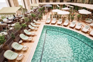 Aerial view of a hotel rooftop pool deck with green cushioned loungers, umbrellas, and seating, overlooking the sea.