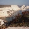 White limestone cliffs along a calm blue coastline, with rocky shore and sparse vegetation in the foreground.