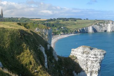 Cliff-top church and grassy fields overlook a blue sea and sandy cove below.