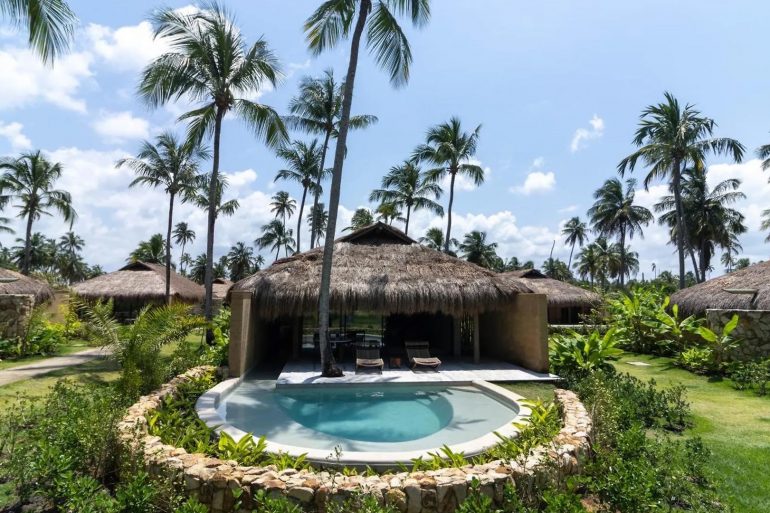 Tropical resort scene with thatched-roof huts, lush palm trees, and a central pool under a blue sky.