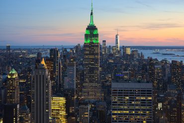 New York City skyline at sunset with the Empire State Building lit green in the center and water in the distance.