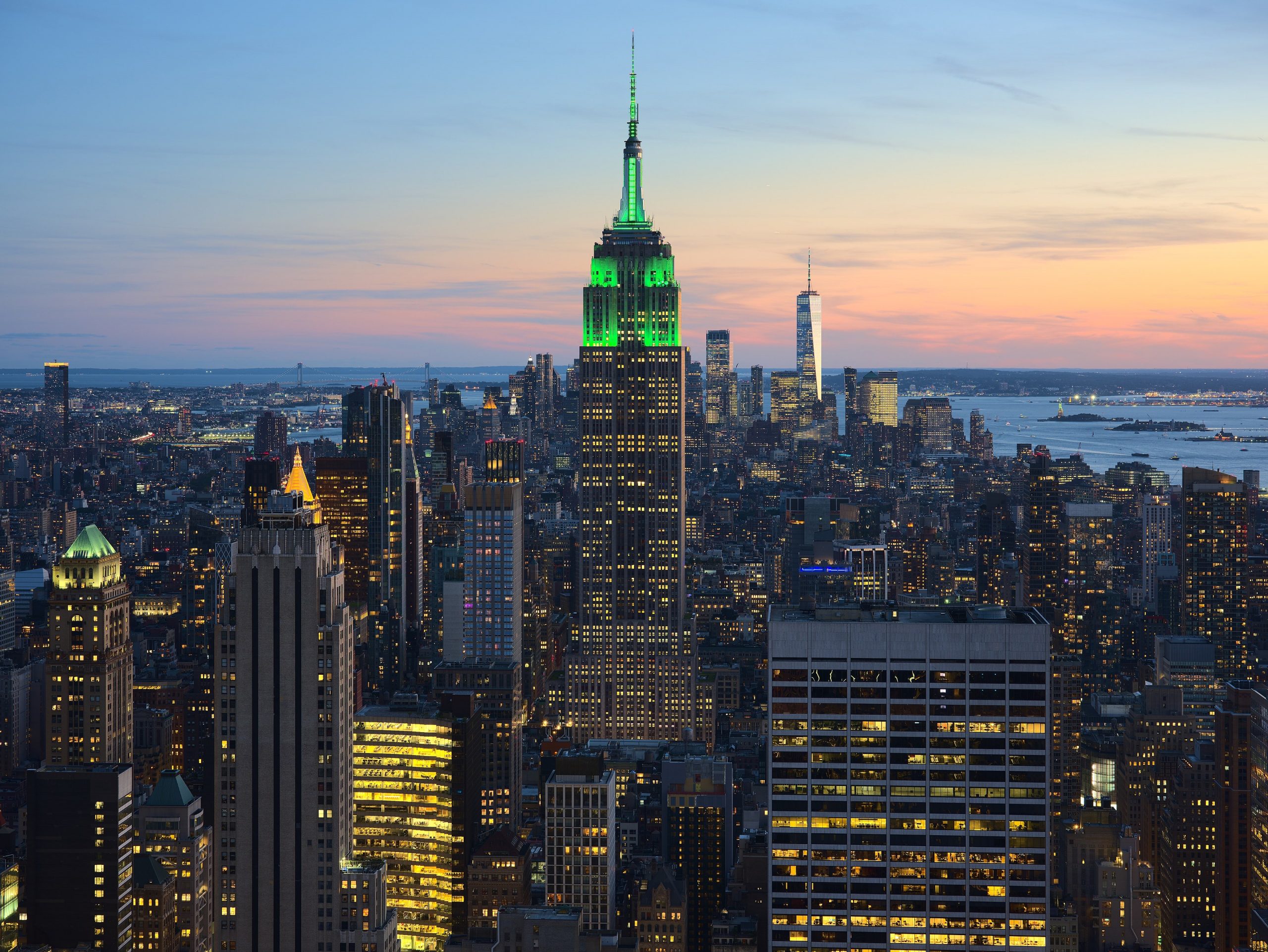 New York City skyline at sunset with the Empire State Building lit green in the center and water in the distance.