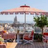 Rooftop cafe with red and white striped umbrellas, orange cushions, and wooden tables overlooking a city skyline with a distant cathedral silhouette.