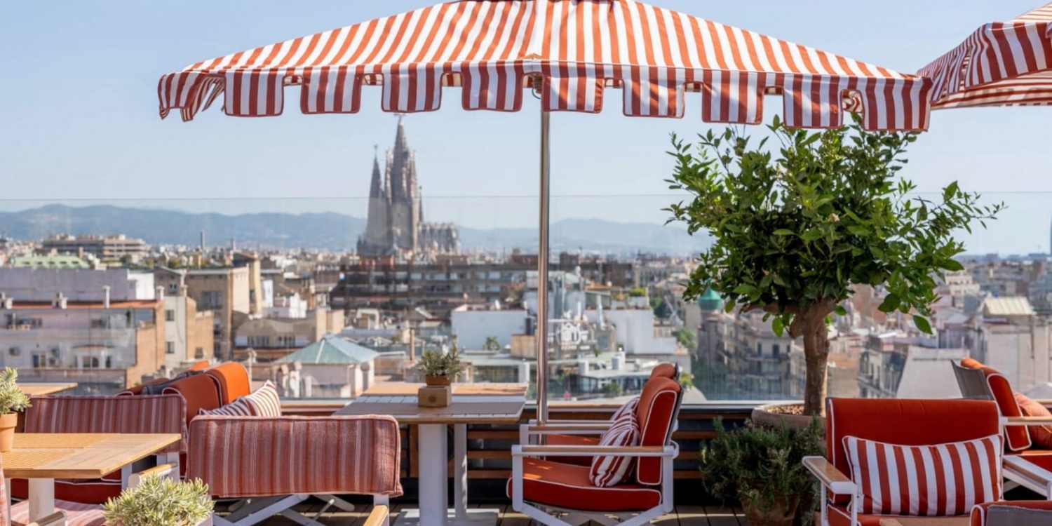 Rooftop cafe with red and white striped umbrellas, orange cushions, and wooden tables overlooking a city skyline with a distant cathedral silhouette.