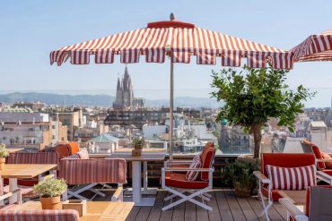 Rooftop cafe with red and white striped umbrellas, orange cushions, and wooden tables overlooking a city skyline with a distant cathedral silhouette.