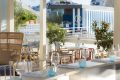 Sunlit outdoor dining terrace with white tables, blue-tinted glassware, and orange trees, overlooking a blue-striped Molitor structure.