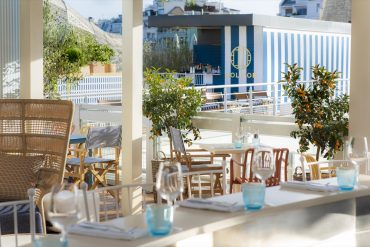Sunlit outdoor dining terrace with white tables, blue-tinted glassware, and orange trees, overlooking a blue-striped Molitor structure.