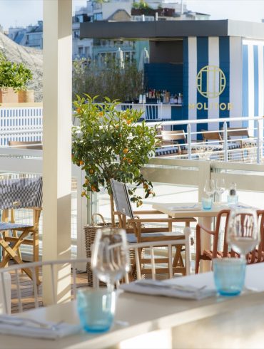 Sunlit outdoor dining terrace with white tables, blue-tinted glassware, and orange trees, overlooking a blue-striped Molitor structure.