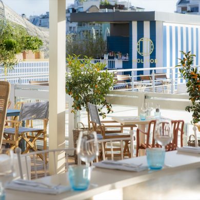 Sunlit outdoor dining terrace with white tables, blue-tinted glassware, and orange trees, overlooking a blue-striped Molitor structure.