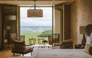 Hotel room with open glass balcony doors revealing a green valley; seating area with wood chairs and small round tables.