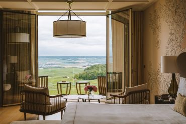Hotel room with open glass balcony doors revealing a green valley; seating area with wood chairs and small round tables.