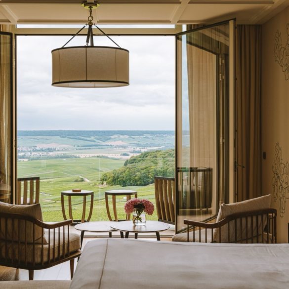 Hotel room with open glass balcony doors revealing a green valley; seating area with wood chairs and small round tables.
