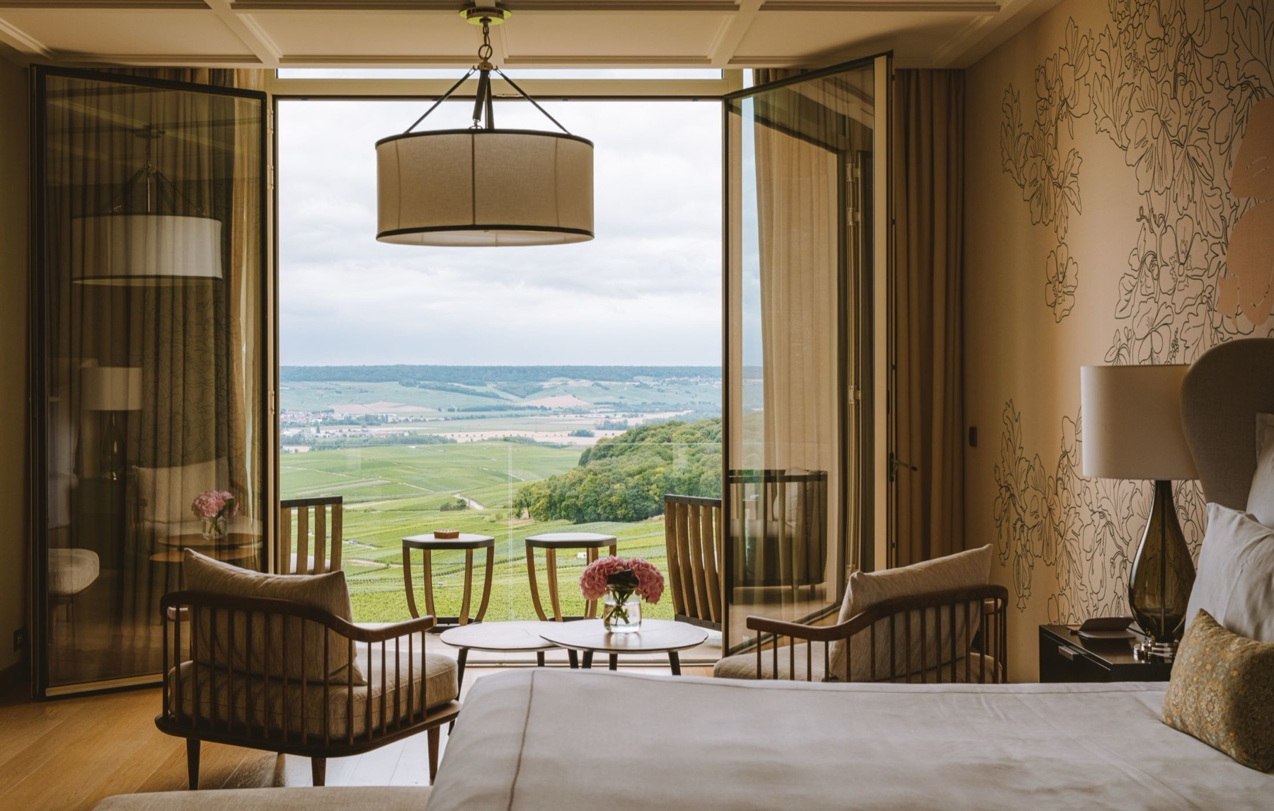 Hotel room with open glass balcony doors revealing a green valley; seating area with wood chairs and small round tables.