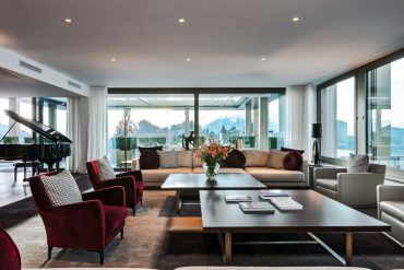 Luxurious living room with a grand piano, beige sectional sofa, red armchairs, and floor-to-ceiling windows showing snowy mountains outside.