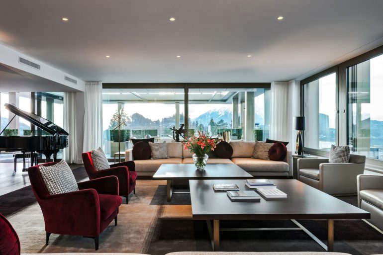 Luxurious living room with a grand piano, beige sectional sofa, red armchairs, and floor-to-ceiling windows showing snowy mountains outside.