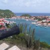 Harbor town with red-roofed houses around a marina of docked boats, hills to the left and the sea in the distance.