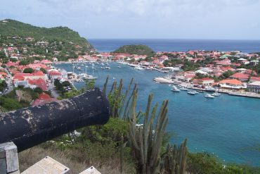 Harbor town with red-roofed houses around a marina of docked boats, hills to the left and the sea in the distance.