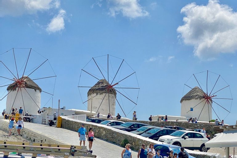 Coastal scene with three white windmills having thatched roofs and large metal sails, under a blue sky with people walking nearby.