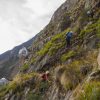 Hikers wearing helmets climb a steep rocky mountainside with glass cable-car pods attached to the cliff.