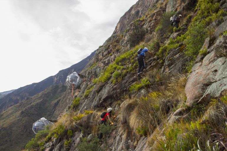 Hikers wearing helmets climb a steep rocky mountainside with glass cable-car pods attached to the cliff.