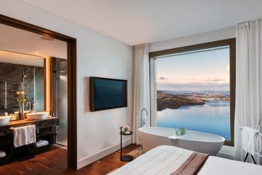Modern hotel room with a freestanding tub by a large window overlooking water and cityscape beyond.