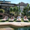 People reclining on poolside lounge chairs by a modern house with tropical plants and a rocky mountain in the background.