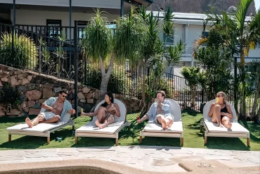 People reclining on poolside lounge chairs by a modern house with tropical plants and a rocky mountain in the background.