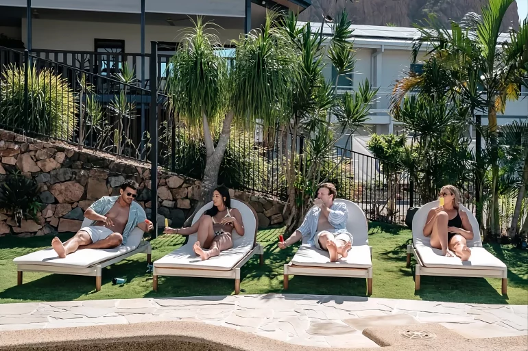 People reclining on poolside lounge chairs by a modern house with tropical plants and a rocky mountain in the background.
