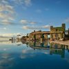 Infinity pool at a luxury resort with ivy-covered stone arches, sun loungers, and a clock tower against a blue sky reflection in the water.