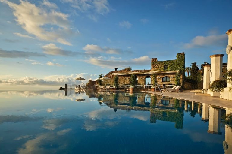 Infinity pool at a luxury resort with ivy-covered stone arches, sun loungers, and a clock tower against a blue sky reflection in the water.