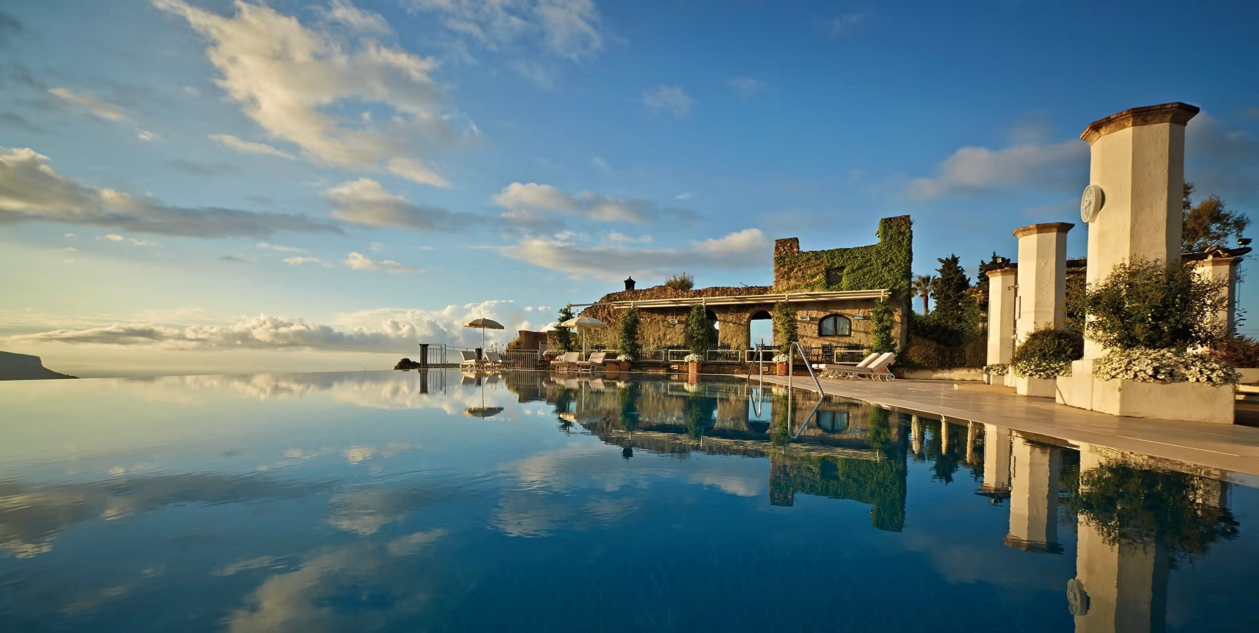 Infinity pool at a luxury resort with ivy-covered stone arches, sun loungers, and a clock tower against a blue sky reflection in the water.