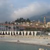 Coastal town with pastel pink buildings, palm-lined promenade, and tall church towers against hills and a cloudy sky.