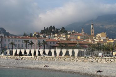 Coastal town with pastel pink buildings, palm-lined promenade, and tall church towers against hills and a cloudy sky.